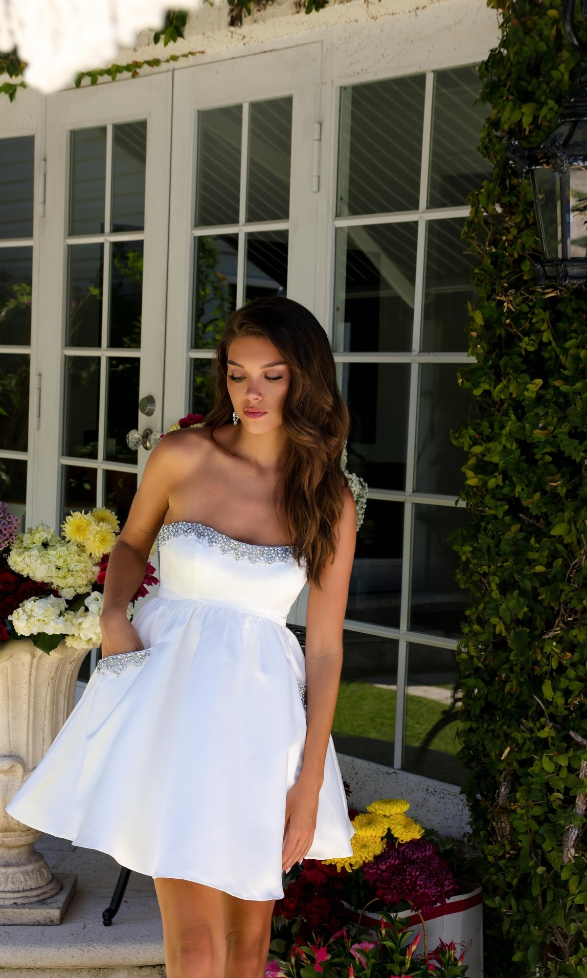 A woman wearing the Ava Presley 29837 strapless short hoco dress with silver trim stands outdoors by a glass door, surrounded by flowers and greenery.